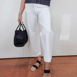 A person wearing white cropped wide-leg pants, a navy patterned short-sleeve top, navy slide sandals, and holding a black bucket bag, standing against a white wall on a terracotta tiled floor.