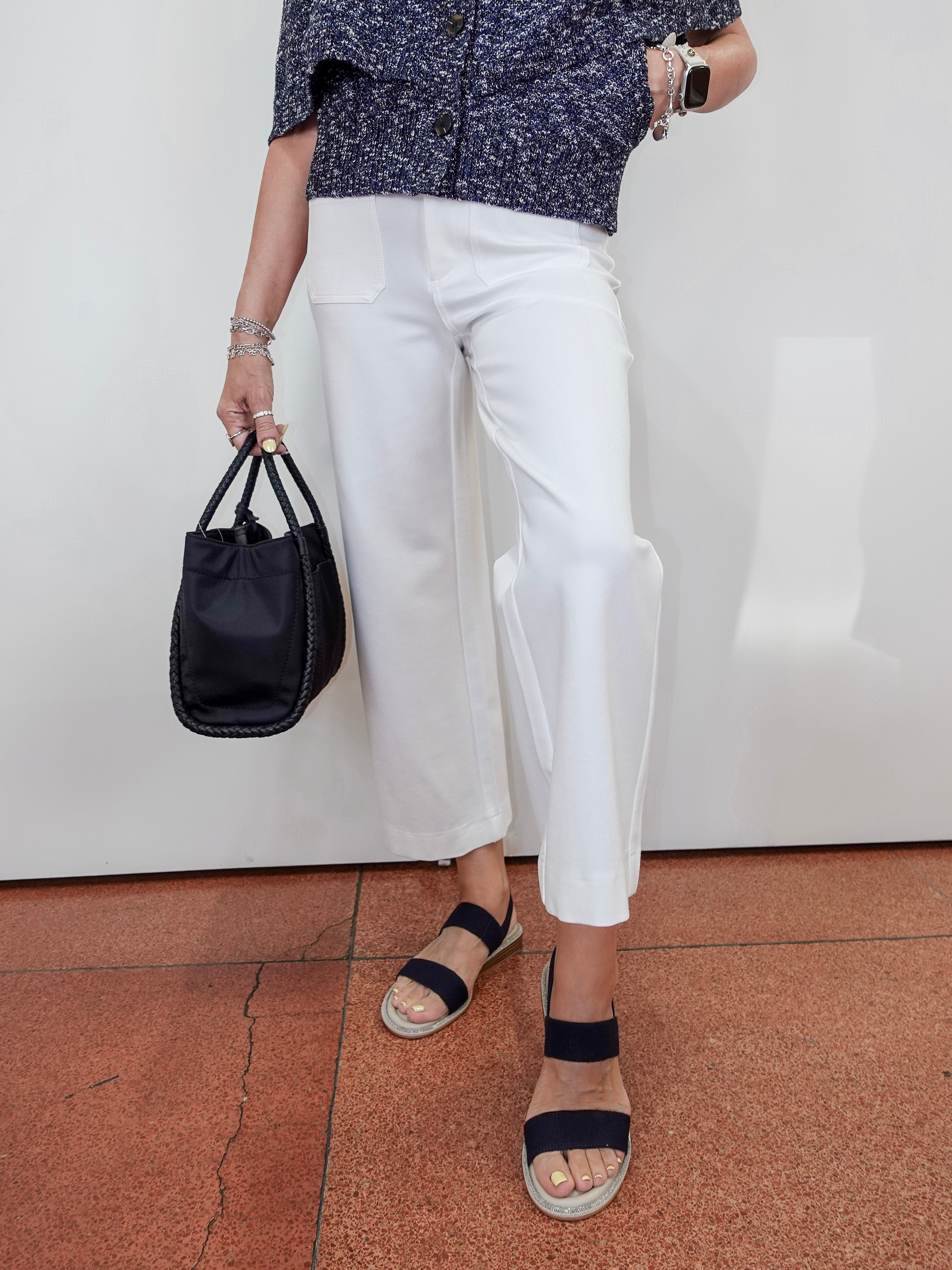 A person wearing white cropped wide-leg pants, a navy patterned short-sleeve top, navy slide sandals, and holding a black bucket bag, standing against a white wall on a terracotta tiled floor.