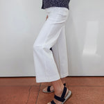 A person wearing white cropped wide-leg pants with a dark patterned top and navy sandals, standing against a white wall on a terracotta tiled floor.