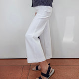 A person wearing white cropped wide-leg pants with a dark patterned top and navy sandals, standing against a white wall on a terracotta tiled floor.