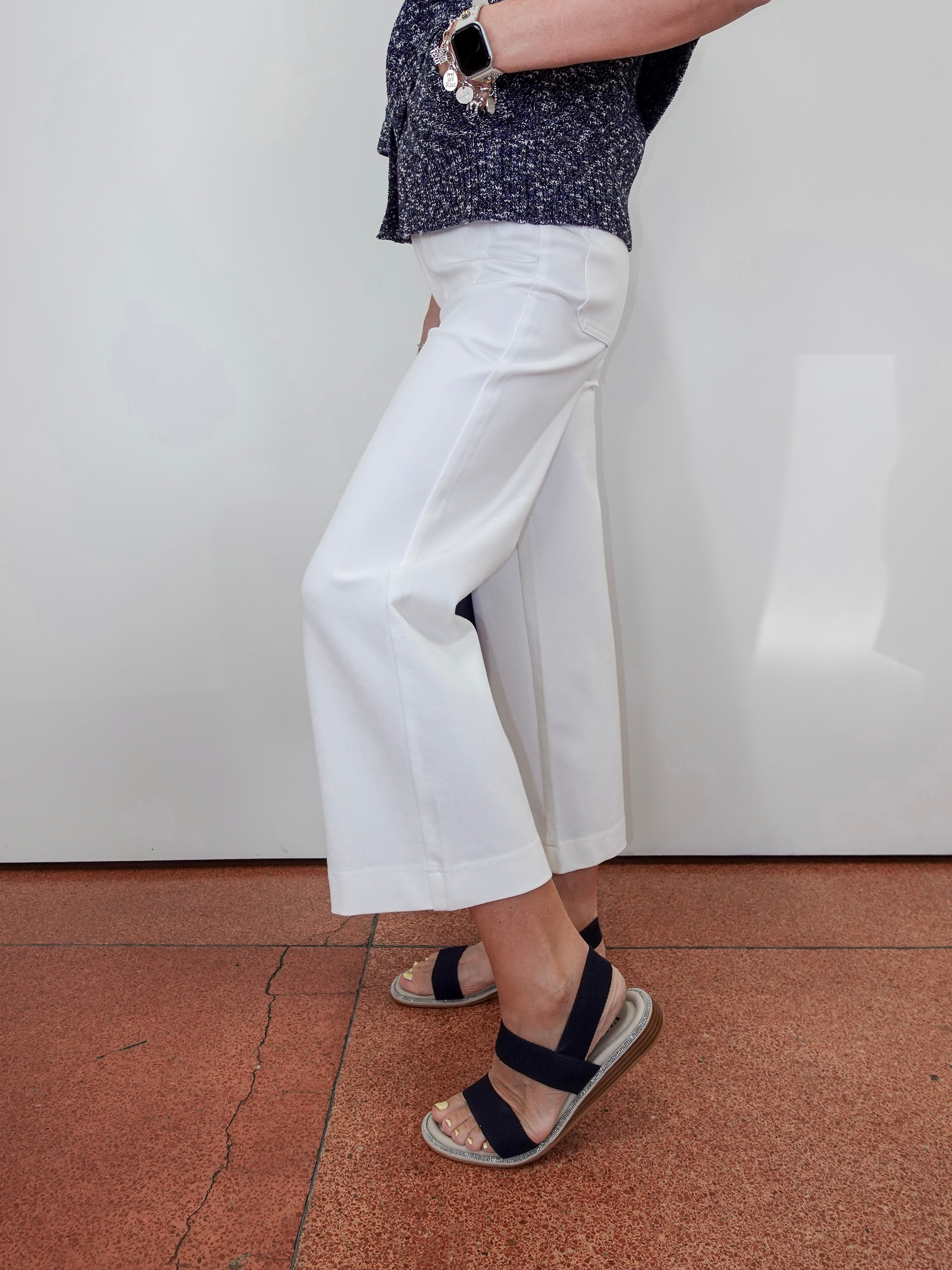 A person wearing white cropped wide-leg pants with a dark patterned top and navy sandals, standing against a white wall on a terracotta tiled floor.