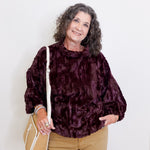 Woman wearing a dark purple textured sweater and beige pants, holding a beige handbag against a white background.