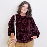 Woman wearing a dark purple textured sweater and beige pants, holding a beige handbag against a white background.