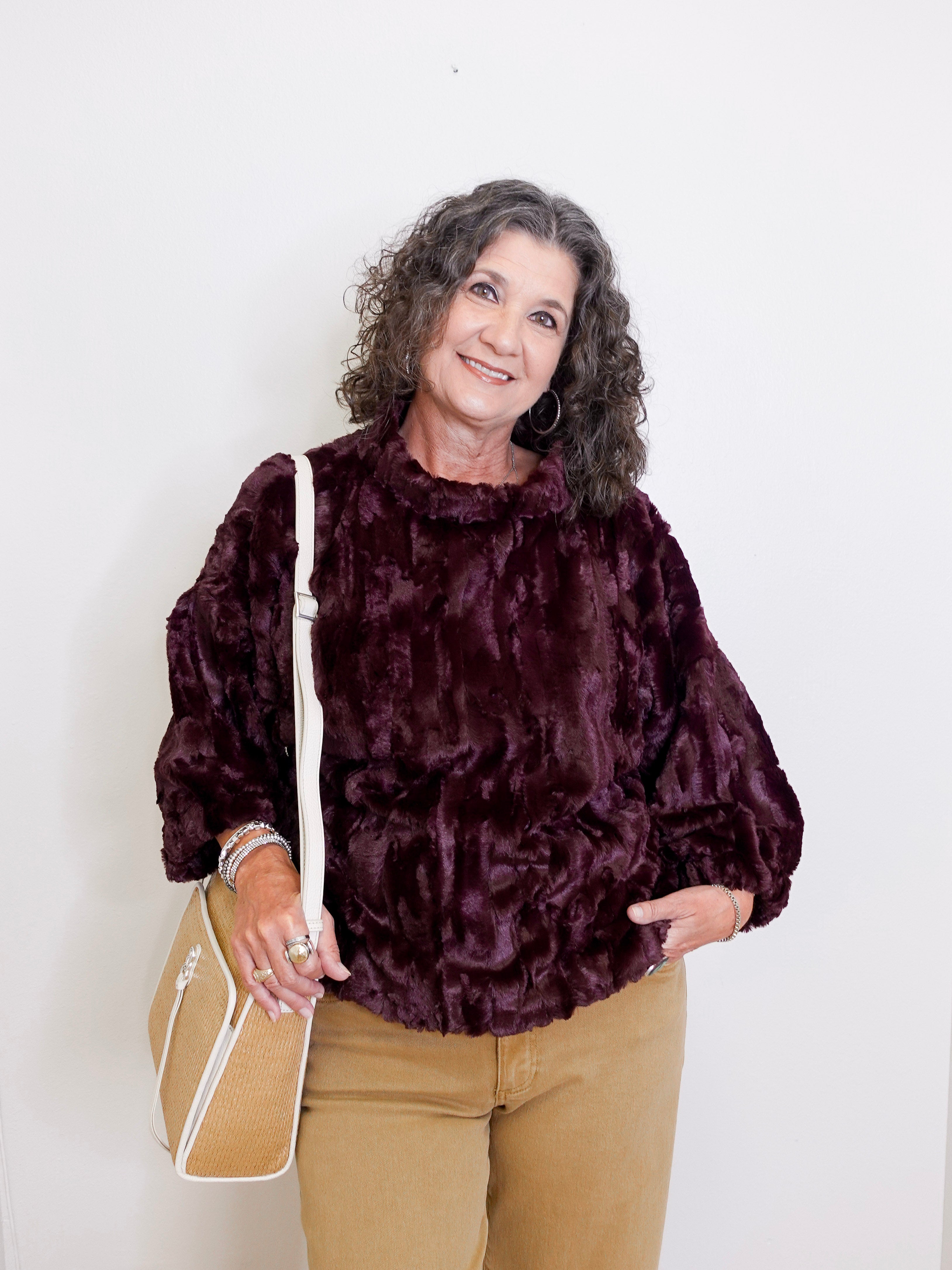 Woman wearing a dark purple textured sweater and beige pants, holding a beige handbag against a white background.