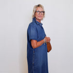 Woman wearing a blue popover shirt dress with pockets in front a white background. 