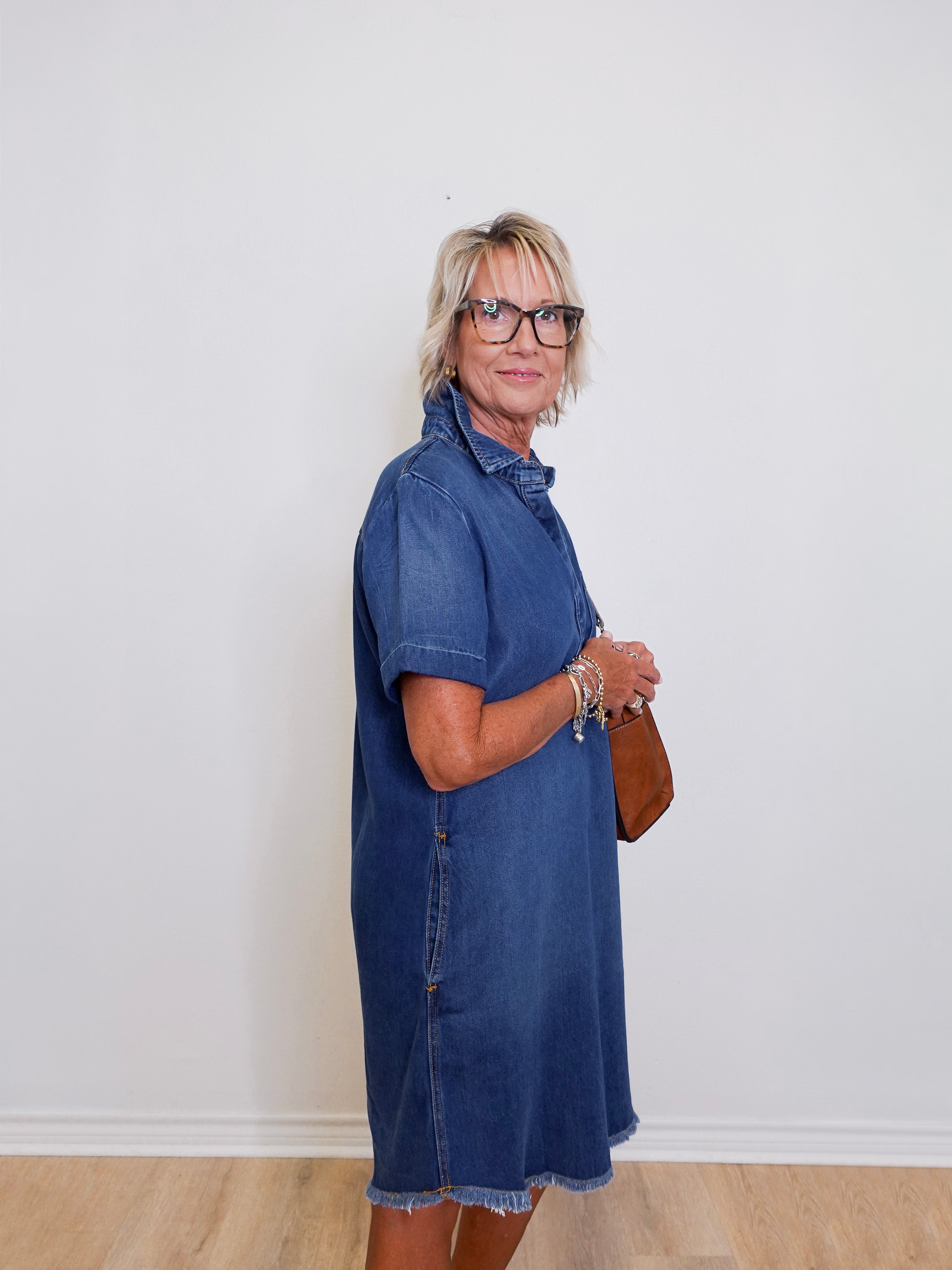 Woman wearing a blue popover shirt dress with pockets in front a white background. 