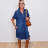 Woman wearing a blue denim dress standing against a white wall.