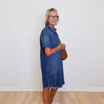 Woman wearing a blue denim dress standing against a white wall.