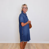 Woman wearing a blue denim dress standing against a white wall.