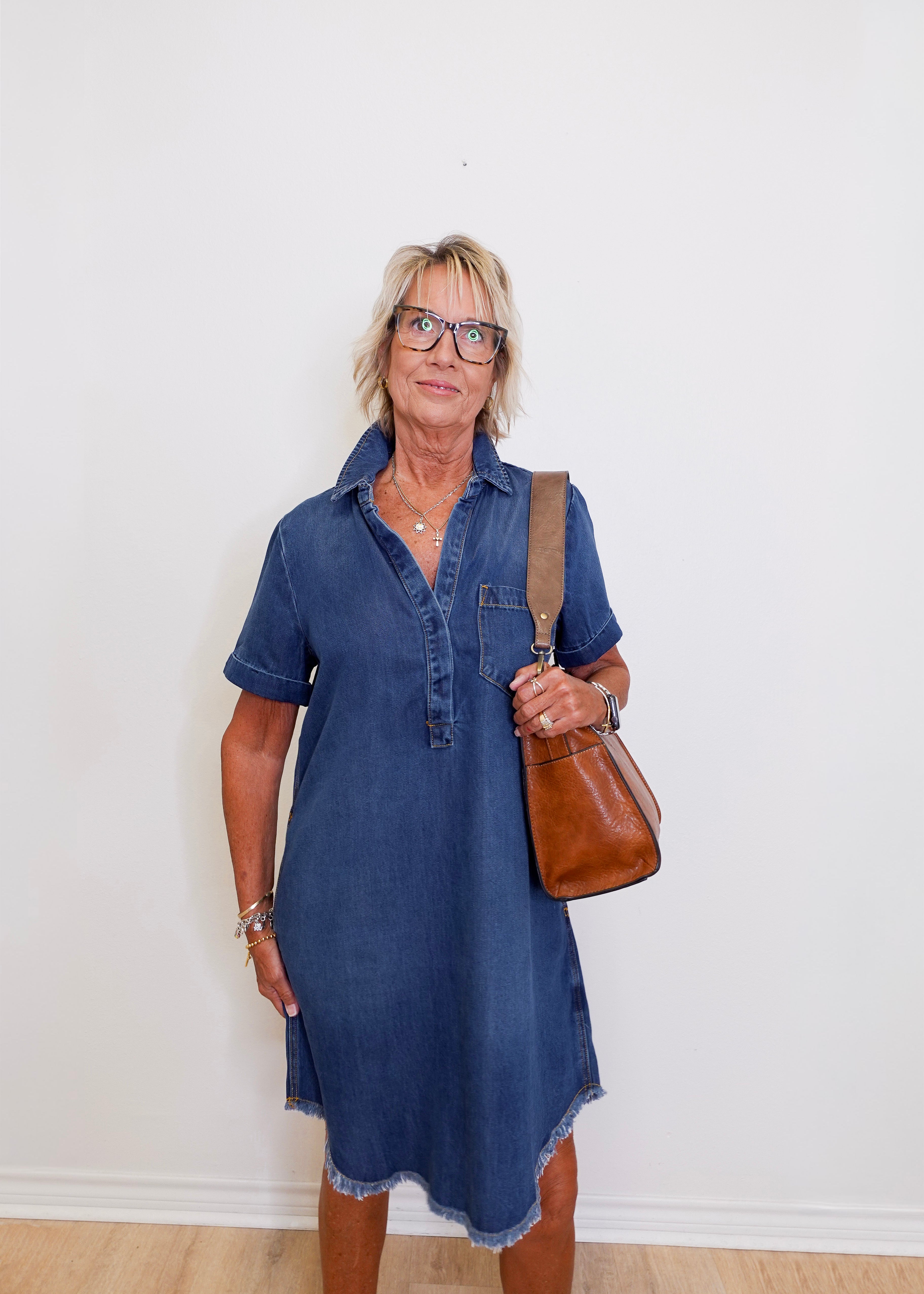 Woman wearing a blue popover shirt dress with pockets in front a white background. 