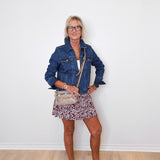 Woman wearing a denim jacket, floral skirt, and white sneakers against a white wall.