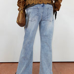 A person shown from the back wearing light-wash blue flare jeans, a brown cropped top with a lace-up back, and carrying a brown shoulder bag against a plain white wall.