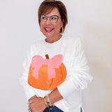 Woman wearing a white sweatshirt with a pumpkin and pink bow design, standing against a plain background.