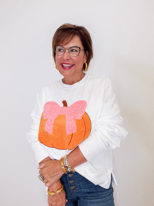 Woman wearing a white sweatshirt with a pumpkin and pink bow design, standing against a plain background.