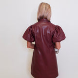 Person wearing a brown leather dress with puffed sleeves against a white wall.