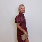 Woman wearing a maroon dress holding a beige handbag against a white wall.