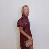 Woman wearing a maroon dress holding a beige handbag against a white wall.