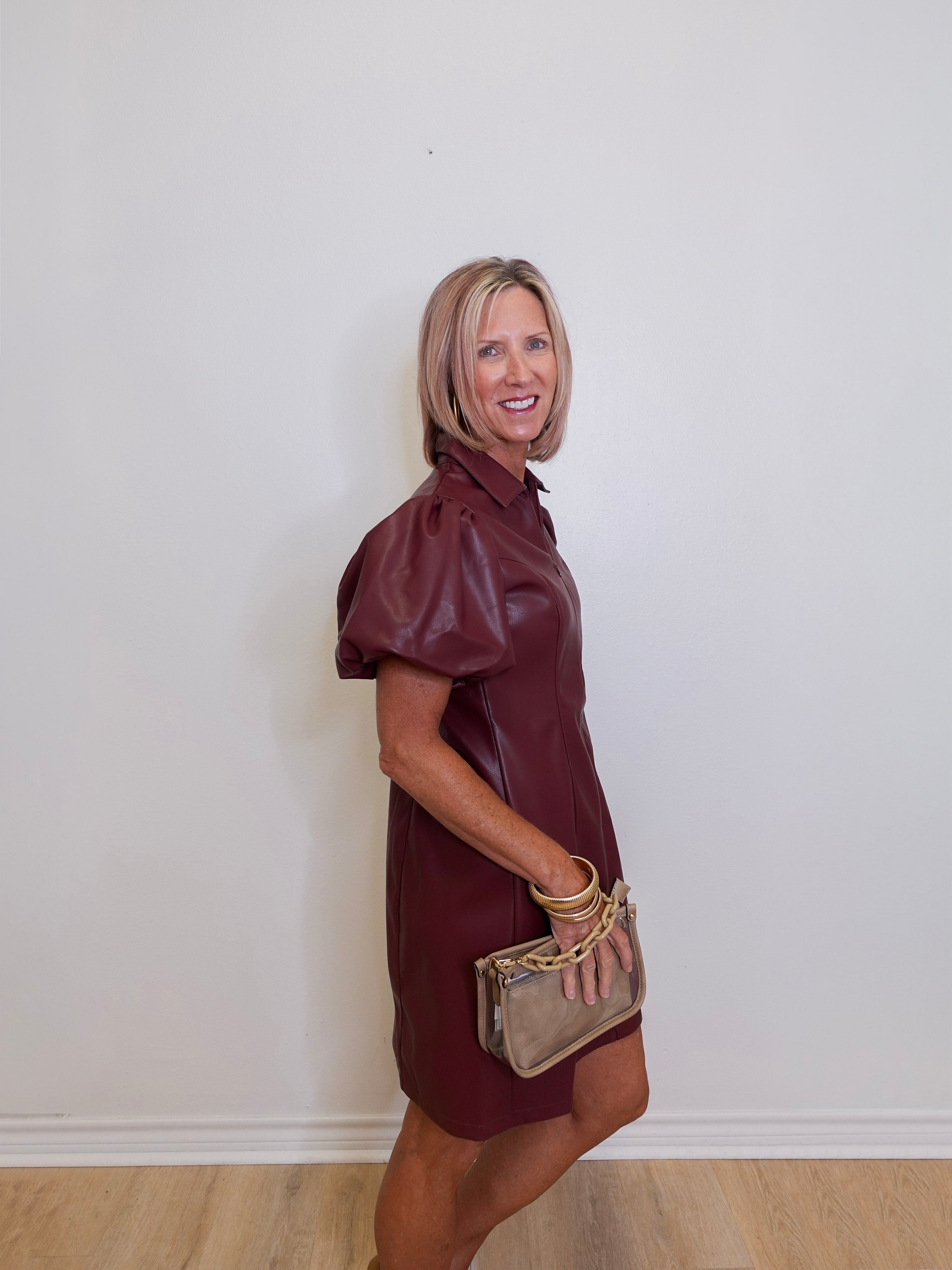 Woman wearing a maroon dress holding a beige handbag against a white wall.