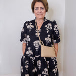 Woman wearing a black floral dress holding a beige handbag against a white wall.