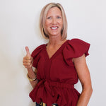 Woman wearing a red blouse with ruffled details, giving a thumbs-up gesture against a plain background.