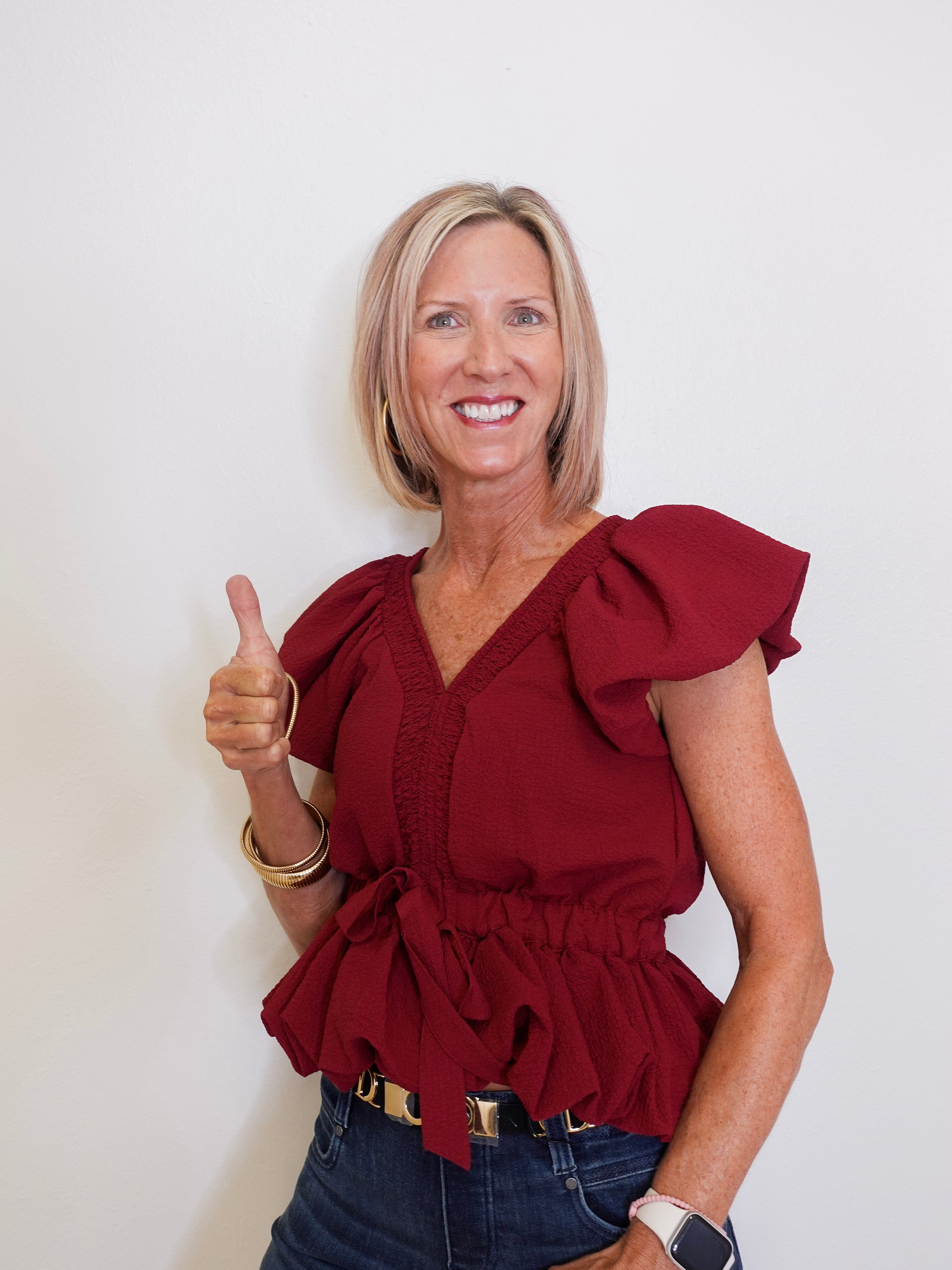 Woman wearing a red blouse with ruffled details, giving a thumbs-up gesture against a plain background.