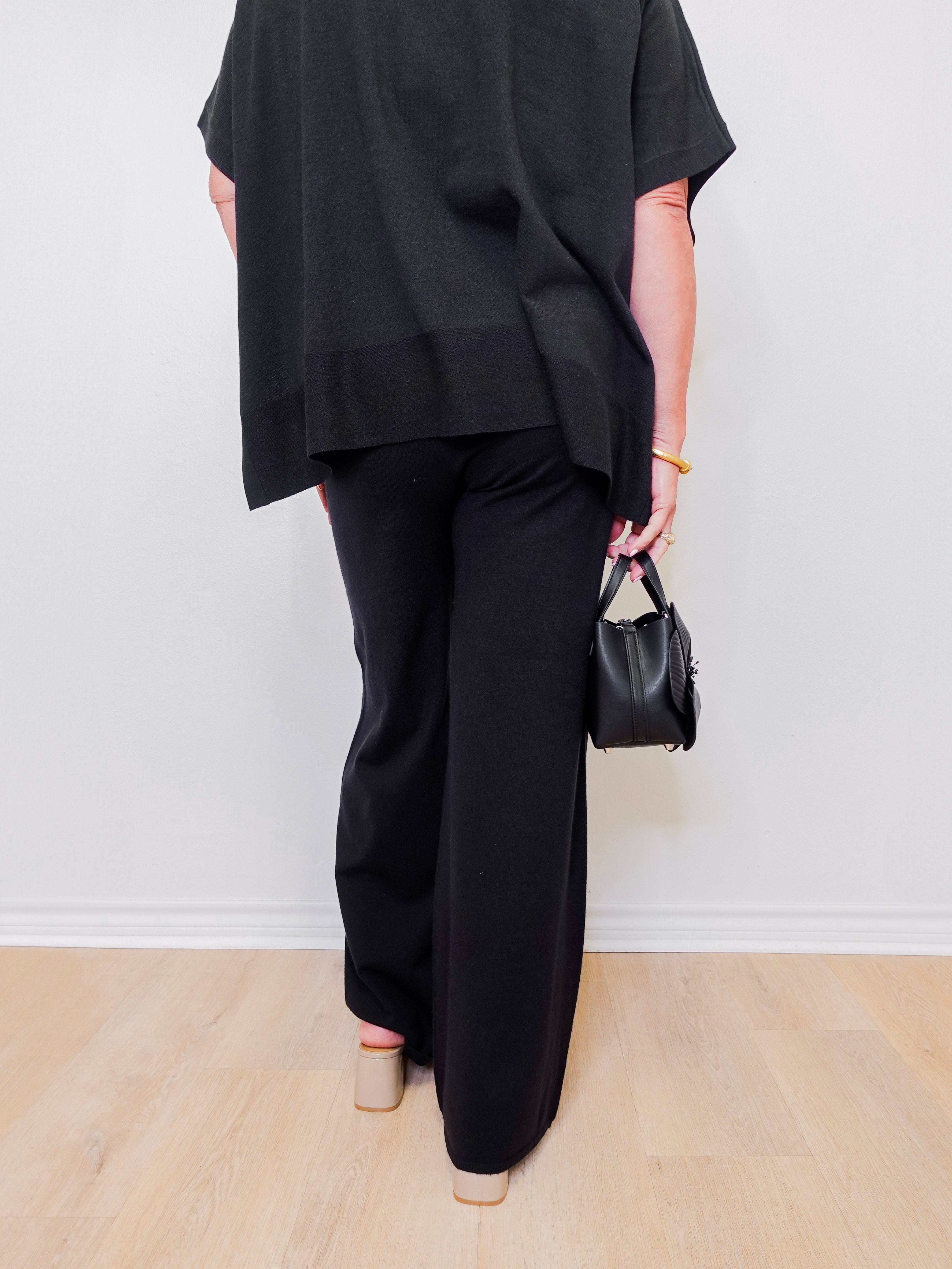 Person wearing a black outfit with wide-leg pants and holding a small black handbag against a white background.