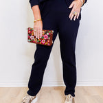 Person wearing black pants and a dark top, holding a floral clutch against a white wall.