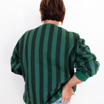 A person shown from the back wearing a dark green and black vertically striped long-sleeve top with light blue jeans, set against a plain white background.