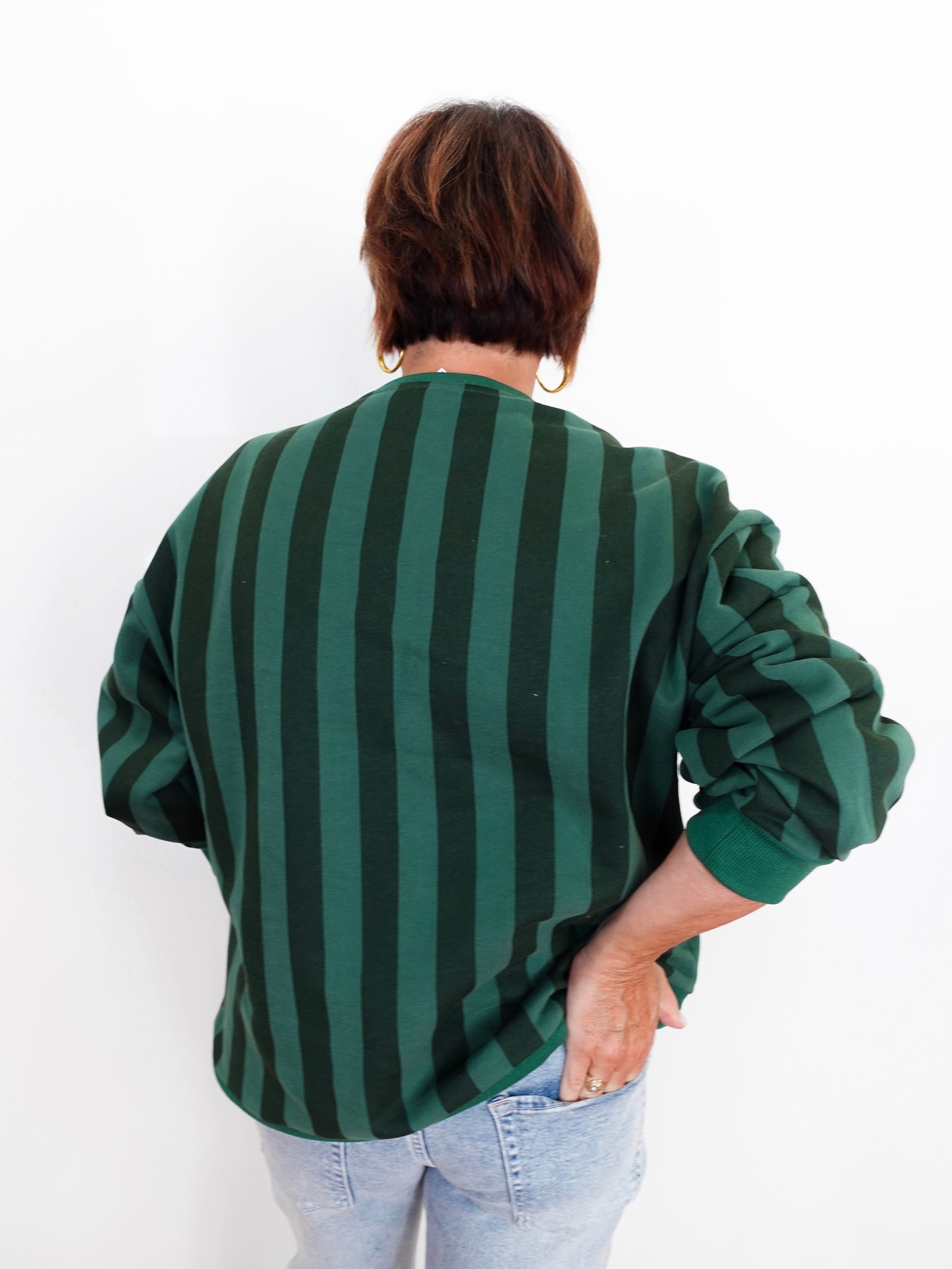 A person shown from the back wearing a dark green and black vertically striped long-sleeve top with light blue jeans, set against a plain white background.