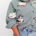 A close-up of a person wearing a green and white gingham button-up shirt with large embroidered ducks across the front and sleeves, paired with light blue jeans against a plain light background.