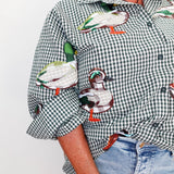 A close-up of a person wearing a green and white gingham button-up shirt with large embroidered ducks across the front and sleeves, paired with light blue jeans against a plain light background.