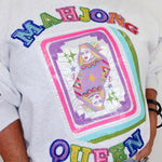 A close-up of a person wearing a white graphic sweatshirt featuring a large sequined 'MAHJONG QUEEN' design on the front, paired with light blue jeans.