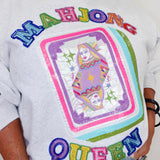 A close-up of a person wearing a white graphic sweatshirt featuring a large sequined 'MAHJONG QUEEN' design on the front, paired with light blue jeans.
