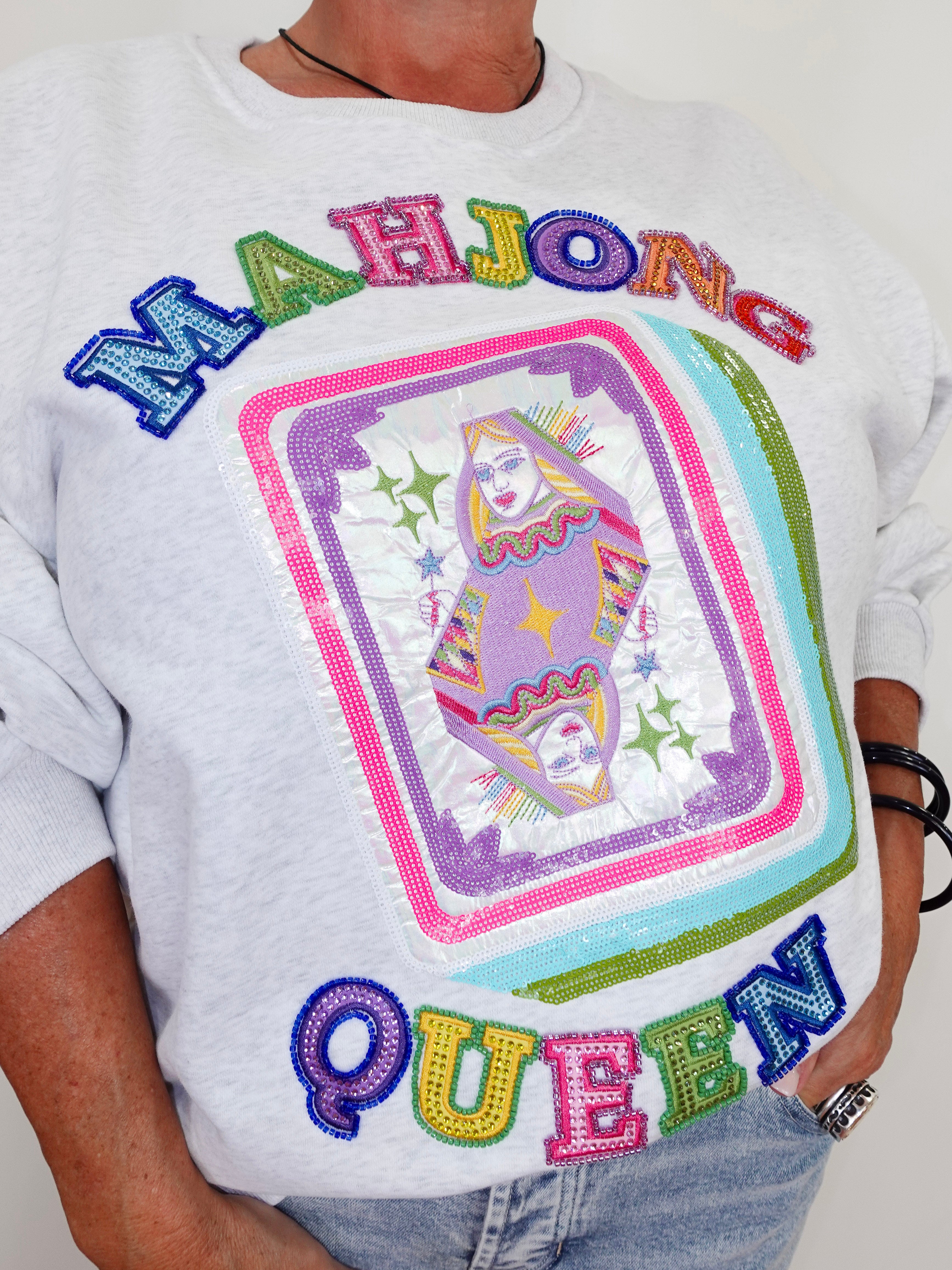 A close-up of a person wearing a white graphic sweatshirt featuring a large sequined 'MAHJONG QUEEN' design on the front, paired with light blue jeans.