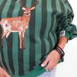 A person wearing a green and dark green vertically striped sweatshirt with a sequined deer applique on the front, paired with light blue jeans. The photo is cropped from the neck down against a plain white background.