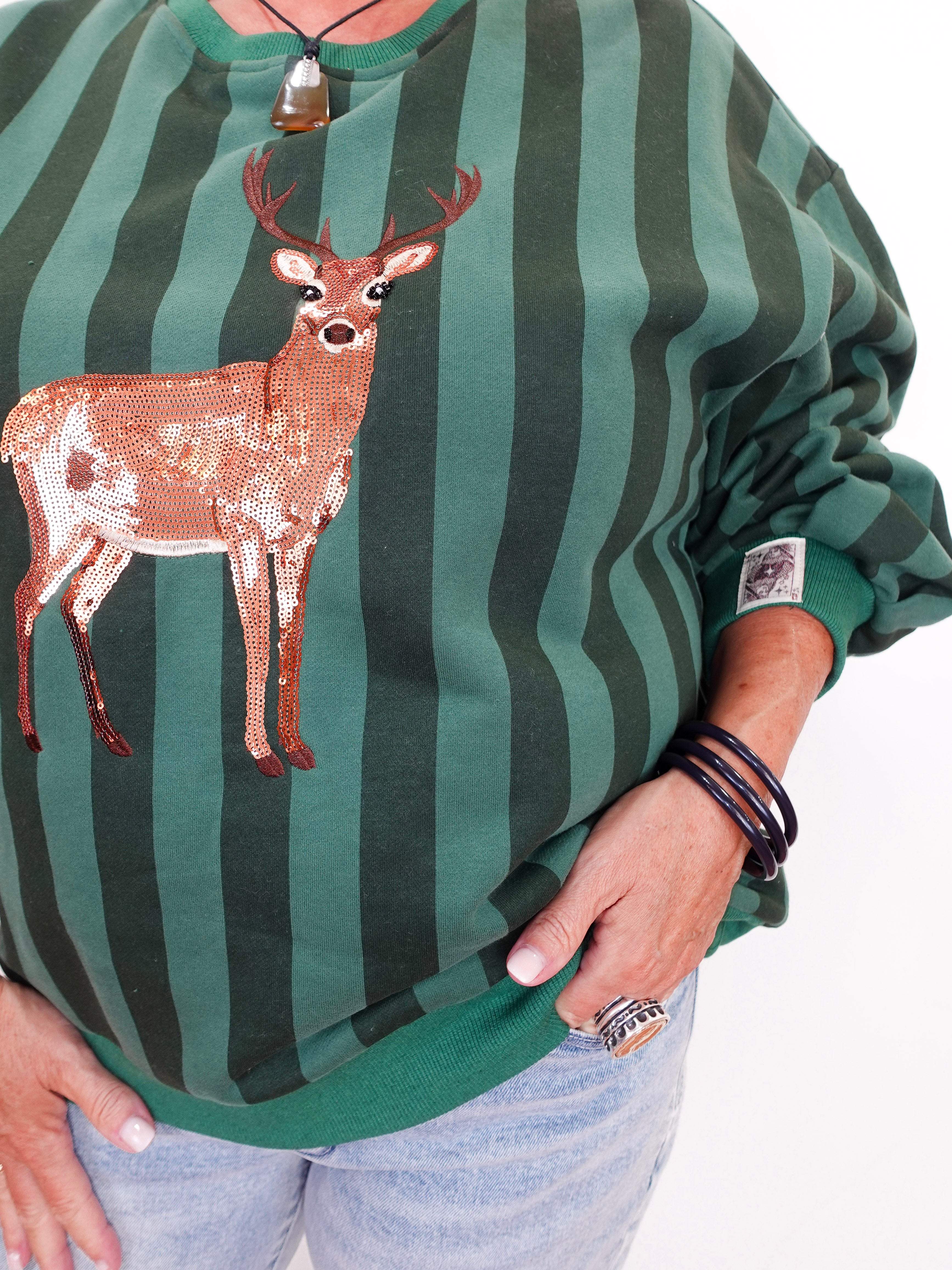 A person wearing a green and dark green vertically striped sweatshirt with a sequined deer applique on the front, paired with light blue jeans. The photo is cropped from the neck down against a plain white background.