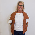 Woman wearing a brown jacket over a white sweater and blue jeans against a white wall.