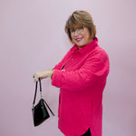 Woman wearing a bright pink jacket holding a black handbag against a light pink background