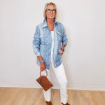 Person wearing a blue denim jacket, white pants, and brown shoes, holding a brown handbag against a white wall.