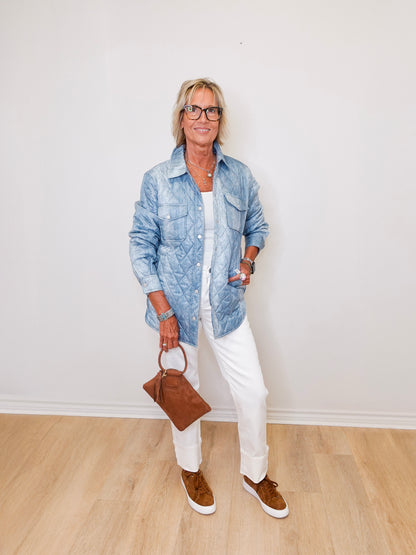 Person wearing a blue denim jacket, white pants, and brown shoes, holding a brown handbag against a white wall.