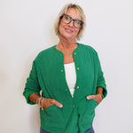 Woman wearing a green cardigan over a white top and blue jeans, standing against a plain background.