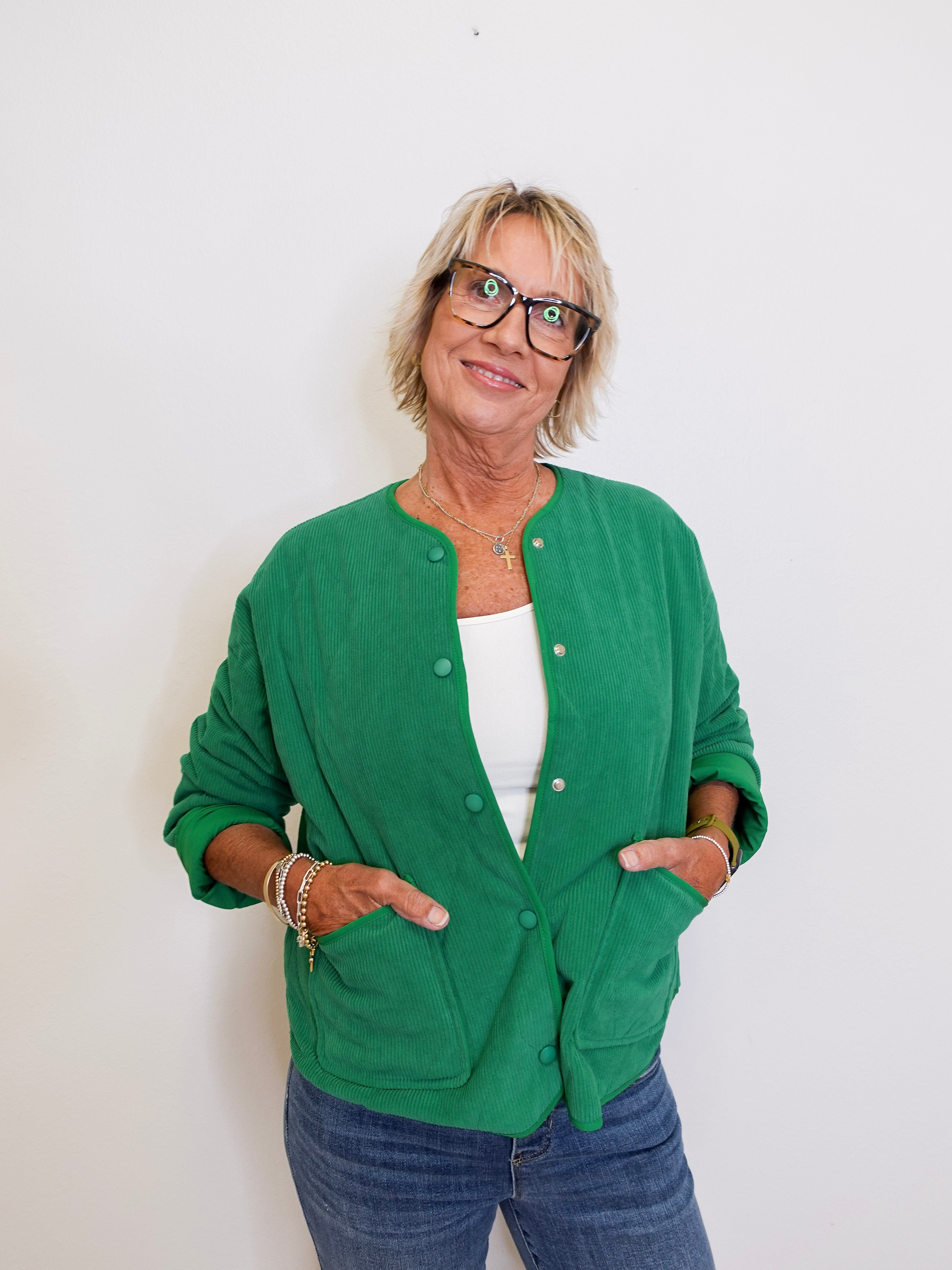 Woman wearing a green cardigan over a white top and blue jeans, standing against a plain background.