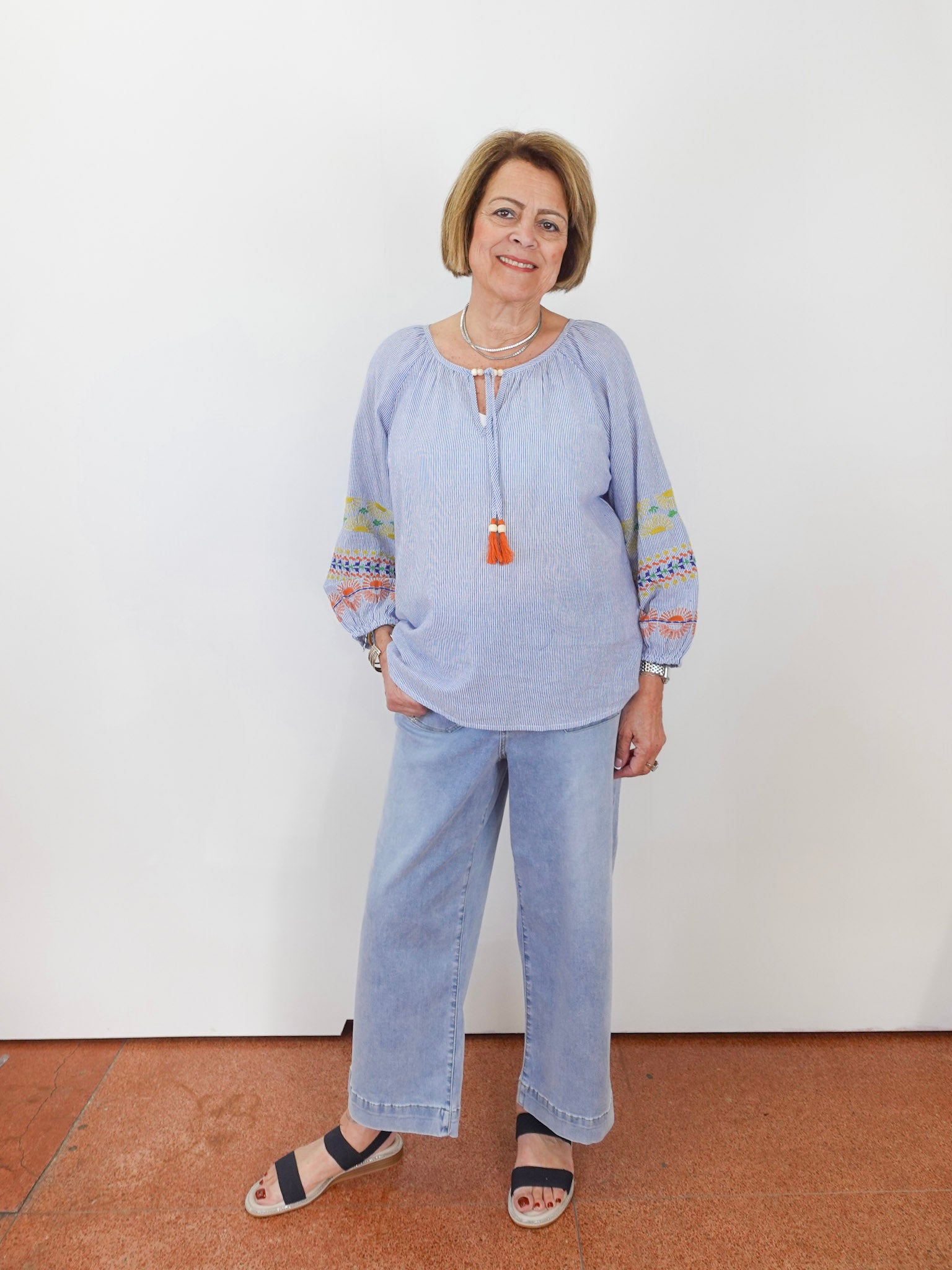 Woman wearing a light blue blouse with colorful sleeves and jeans against a white wall.