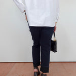 A person shown from the back wearing an oversized white long-sleeve shirt, dark navy cropped pants, and black block-heel sandals, holding a small black handbag against a plain white wall and terracotta floor.
