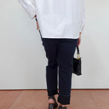 A person shown from the back wearing an oversized white long-sleeve shirt, dark navy cropped pants, and black block-heel sandals, holding a small black handbag against a plain white wall and terracotta floor.