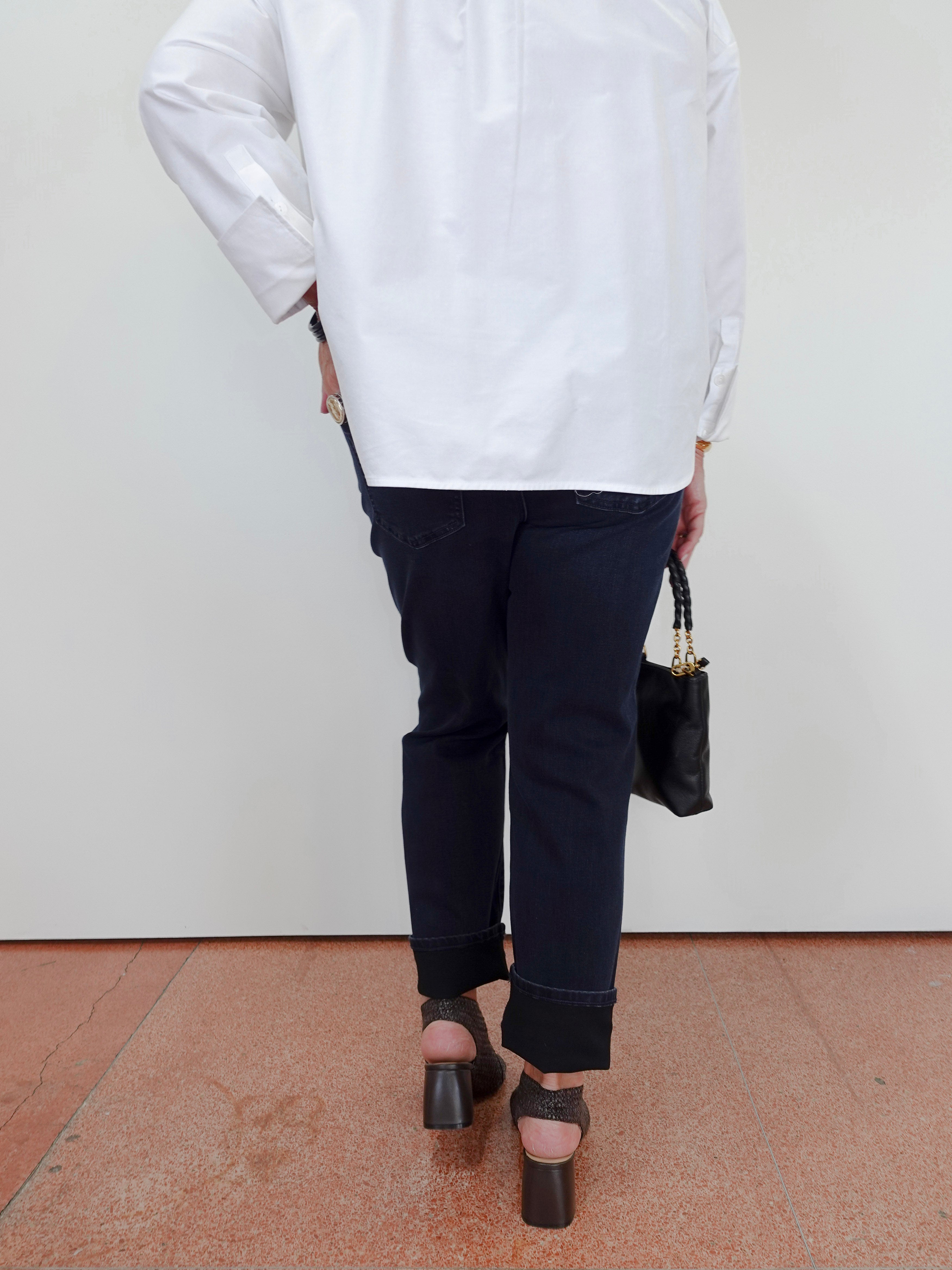 A person shown from the back wearing an oversized white long-sleeve shirt, dark navy cropped pants, and black block-heel sandals, holding a small black handbag against a plain white wall and terracotta floor.