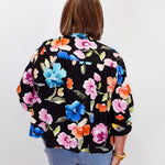 A woman viewed from behind wearing a black long-sleeve blouse with a vibrant multicolor floral print in pink, blue, orange, and green, paired with light blue jeans against a plain white background.