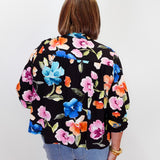 A woman viewed from behind wearing a black long-sleeve blouse with a vibrant multicolor floral print in pink, blue, orange, and green, paired with light blue jeans against a plain white background.