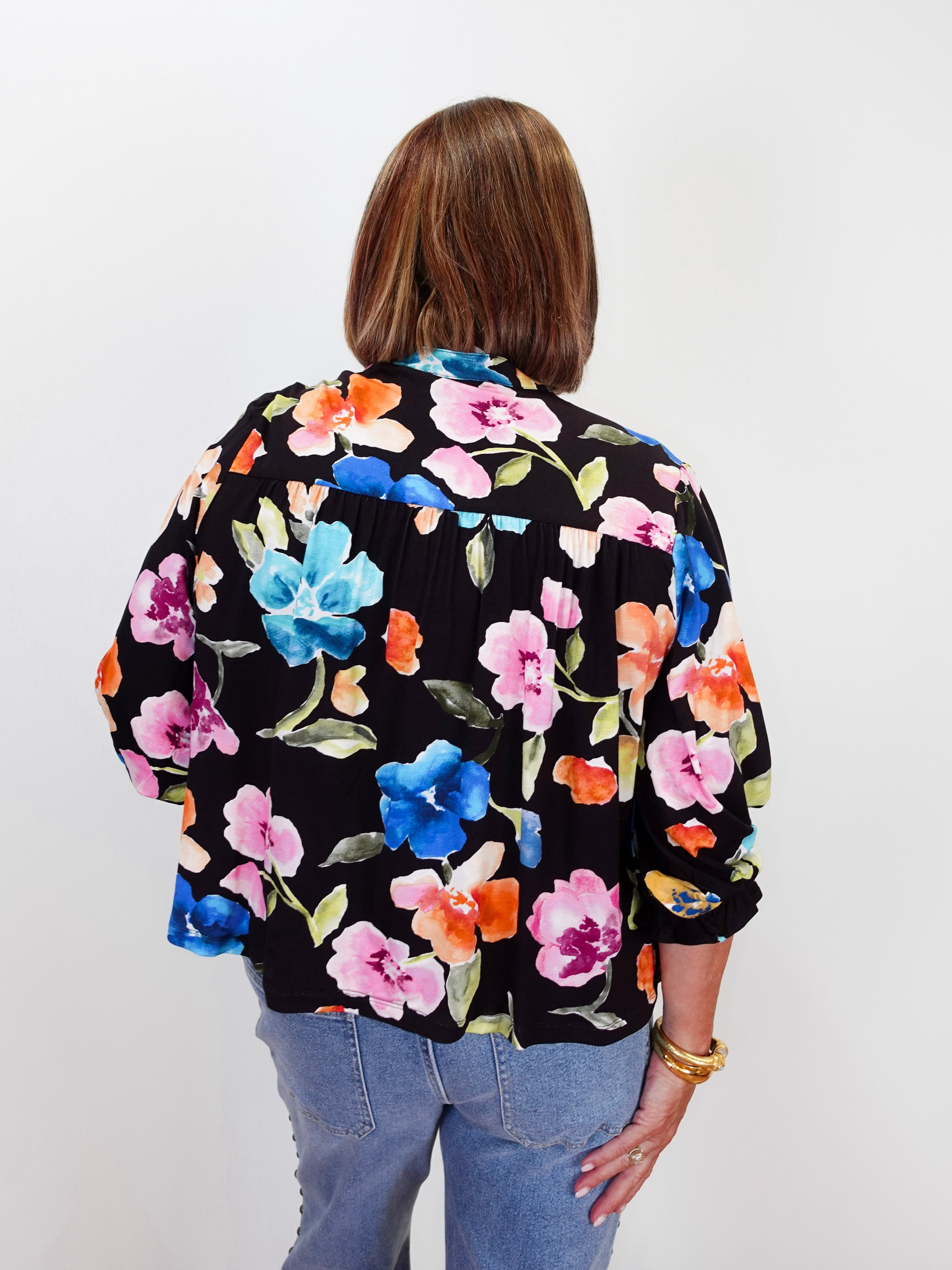 A woman viewed from behind wearing a black long-sleeve blouse with a vibrant multicolor floral print in pink, blue, orange, and green, paired with light blue jeans against a plain white background.