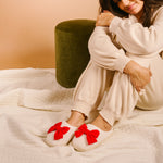 Person wearing white slippers with red bows sitting on a bed.
